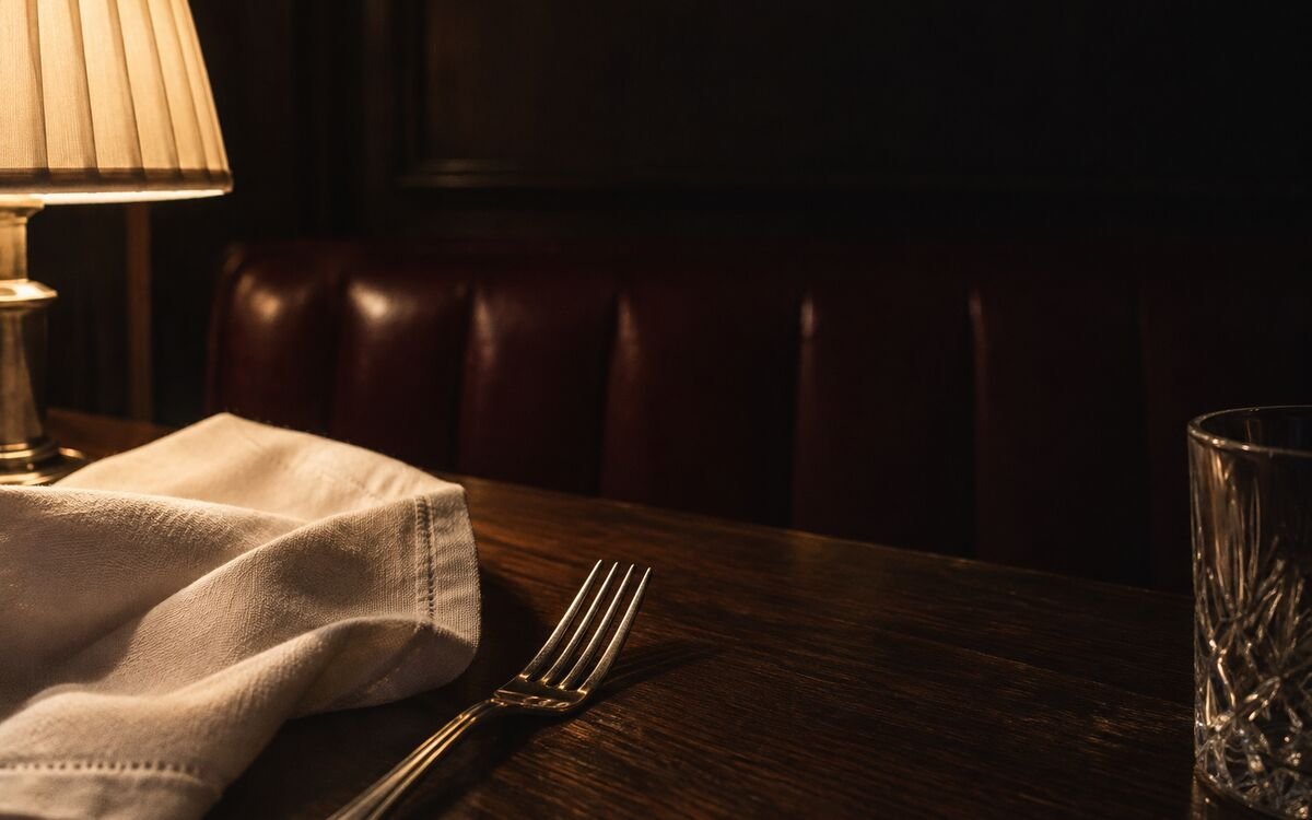 Lamplight on linen and silver beside a dark leather booth